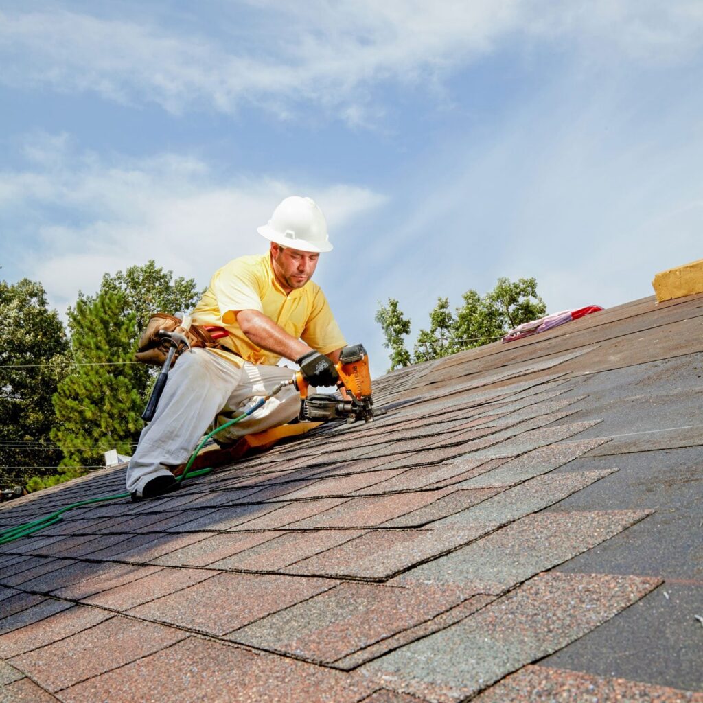 Roofer installing shingles on a residential roof under a clear blue sky.