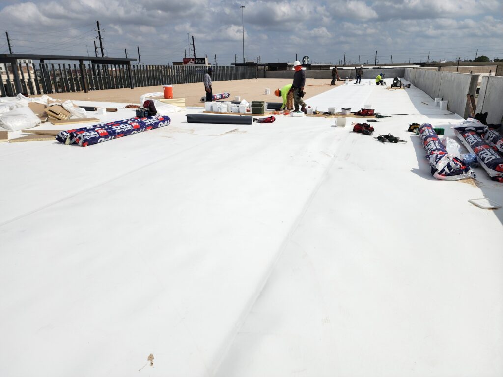 Workers installing roofing material on a flat rooftop under a cloudy sky.